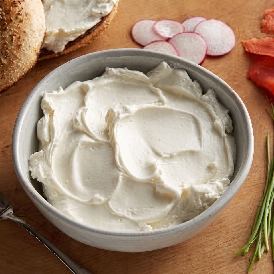 Bowl of creamy white dip surrounded by seeded bread, radish slices, tomatoes, and green onions on a wooden board.