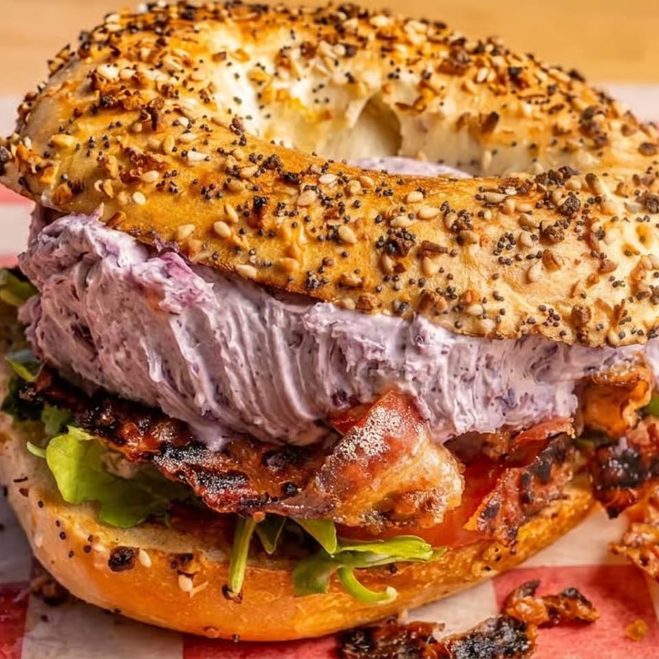 Loaded everything bagel sandwich with purple cream cheese, bacon, greens, and tomato on red-and-white checkered paper.