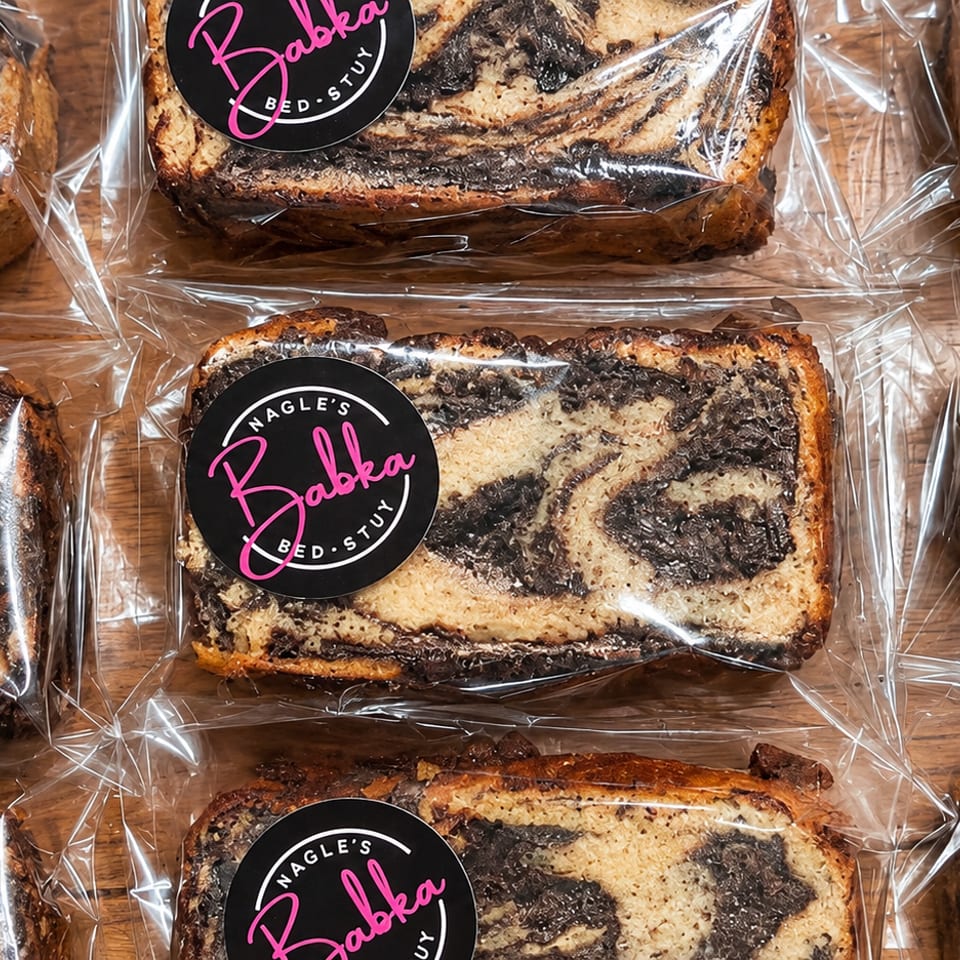 Packaged Babka loaves with swirled chocolate filling arranged across a wooden surface.
