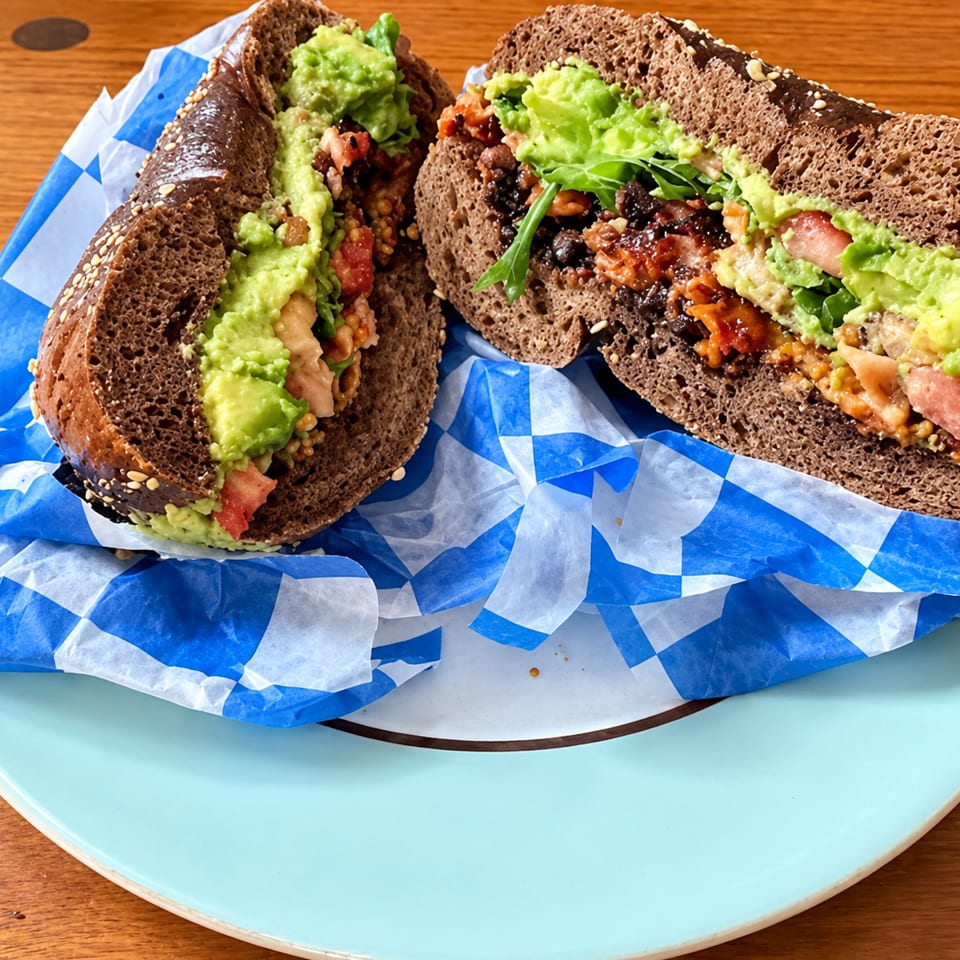 Two dark bread sandwiches filled with mashed avocado, bacon, beans, vegetables, and greens on blue-checkered paper.